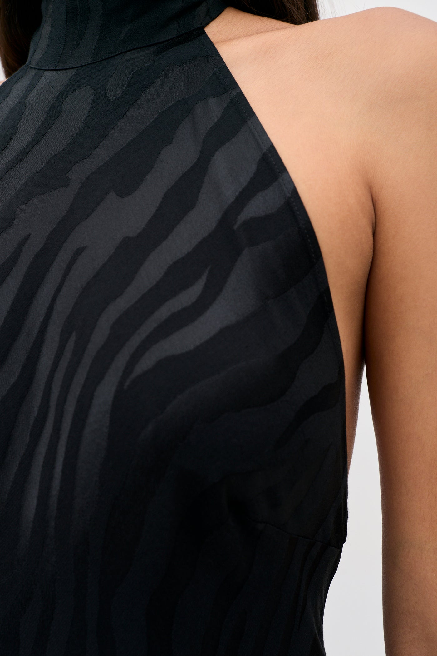 Close-up of the Maura - Cooper Jacquard Black dress, a sleeveless halter style featuring a subtle zebra print pattern, showing part of a person’s shoulder and upper chest against a plain background.