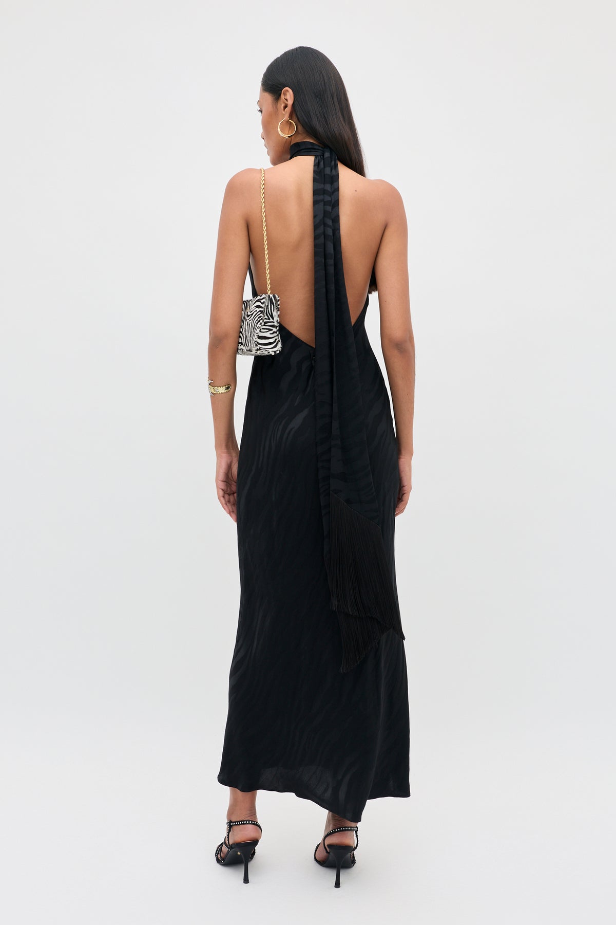 Woman wearing the Maura - Cooper Jacquard Black dress, a long, backless black halter style, paired with a zebra-patterned purse over her shoulder. She stands against a plain white background with straight hair worn down, accessorized with high heels and a bracelet.