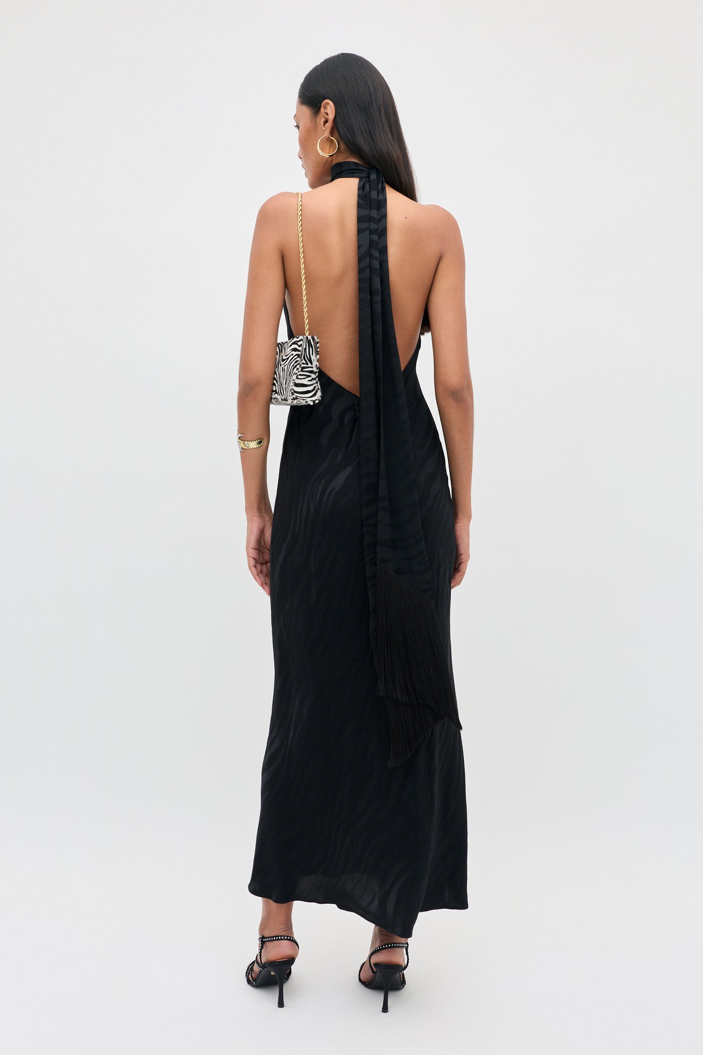 Woman wearing the Maura - Cooper Jacquard Black dress, a long, backless black halter style, paired with a zebra-patterned purse over her shoulder. She stands against a plain white background with straight hair worn down, accessorized with high heels and a bracelet.