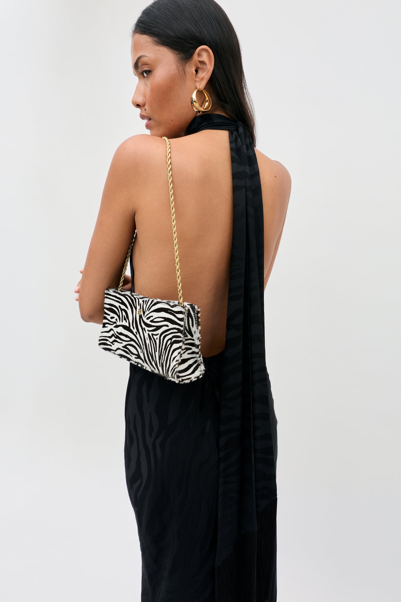 A woman stands sideways in the Maura - Cooper Jacquard Black dress, holding a zebra-print purse with a gold chain strap over her shoulder and wearing large gold hoop earrings.