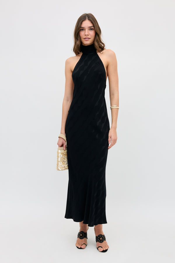 A woman with loose wavy hair stands against a plain background, wearing the Maura - Wave black halter-neck ankle-length dress. She carries a woven handbag, wears black sandals, and accessorizes with a bracelet and ring.
