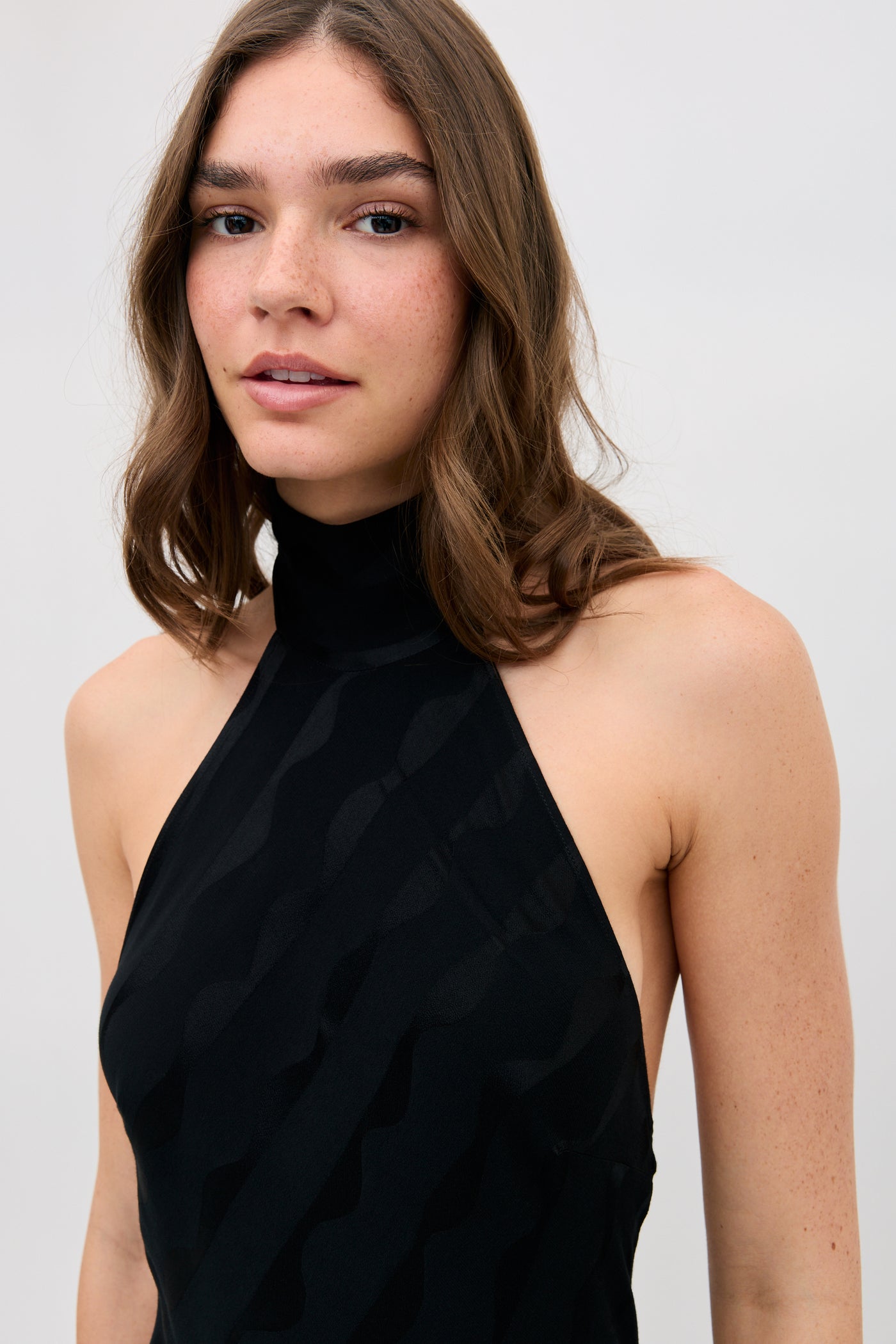 A woman with wavy brown hair and freckles wears the Maura - Wave sleeveless black halter-neck top, standing against a plain light background with a neutral expression while facing the camera.