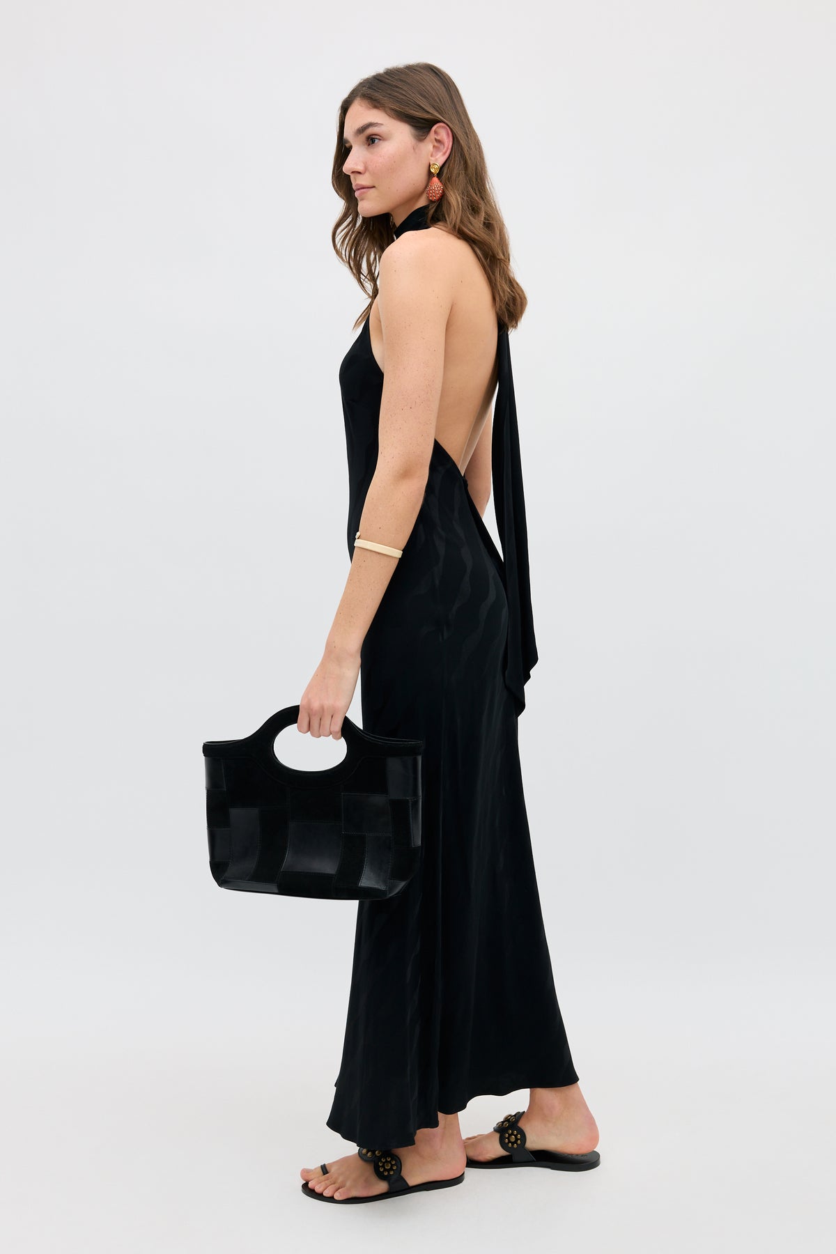 A woman wears the Maura - Wave, a sleeveless, backless black dress with slide sandals. She holds a black handbag and accessorizes with hoop earrings and a gold bracelet, standing against a plain light background.