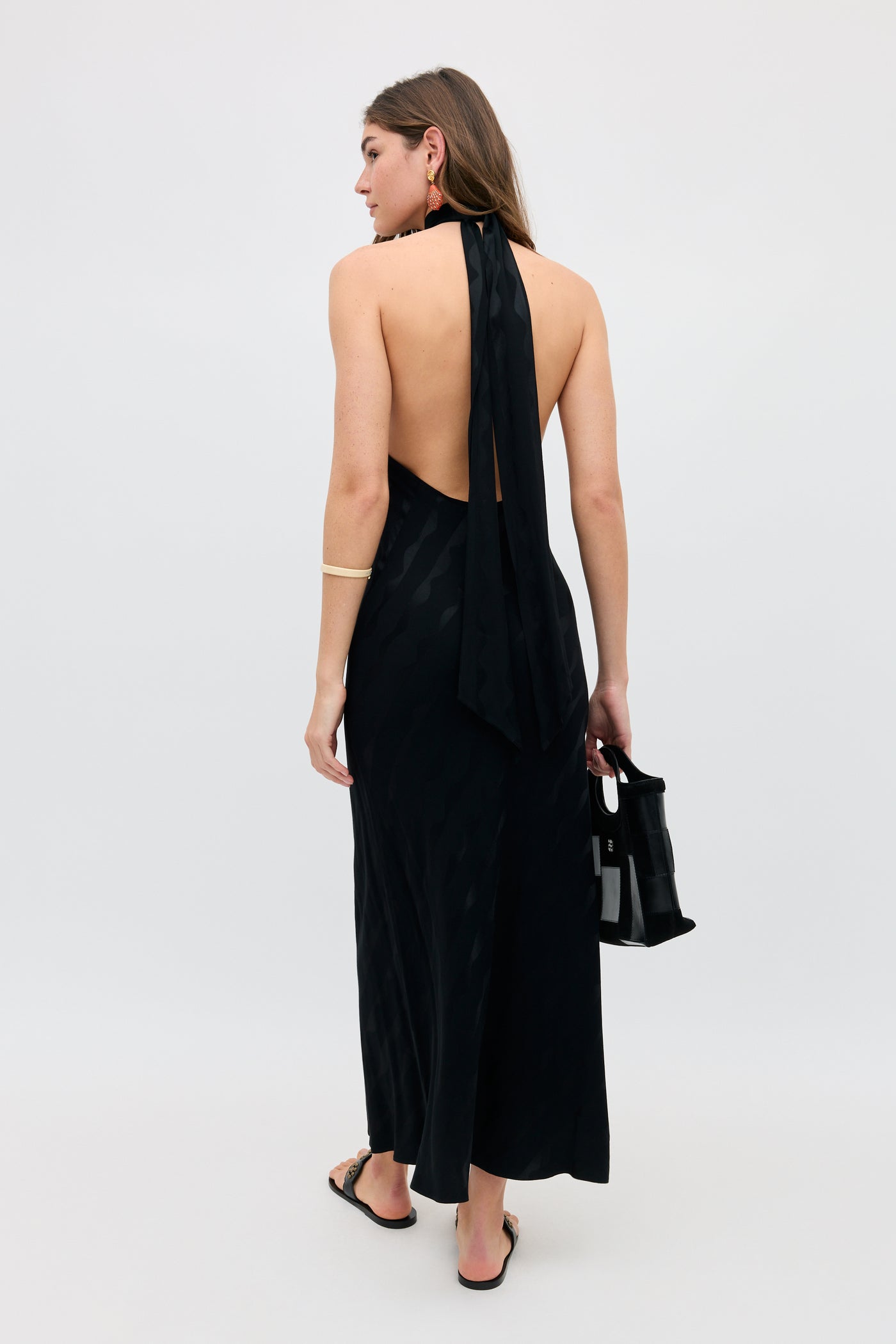 A woman faces away in the Maura - Wave long black halter-neck dress with an open back, holding a small black handbag and wearing hoop earrings, a bracelet, and sandals. The background is plain and light-colored.