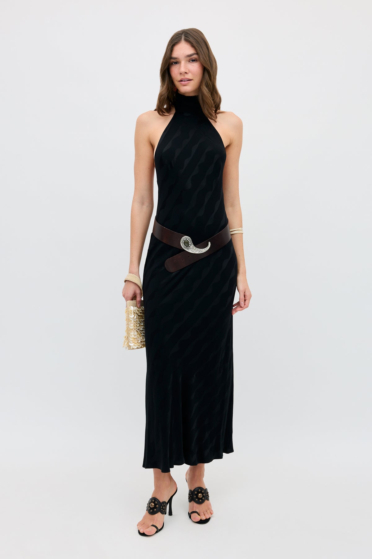 Wearing the Maura - Wave sleeveless black halter dress with a brown belt, black heeled sandals, gold bracelets, and a gold textured handbag, a woman stands against a plain white background.