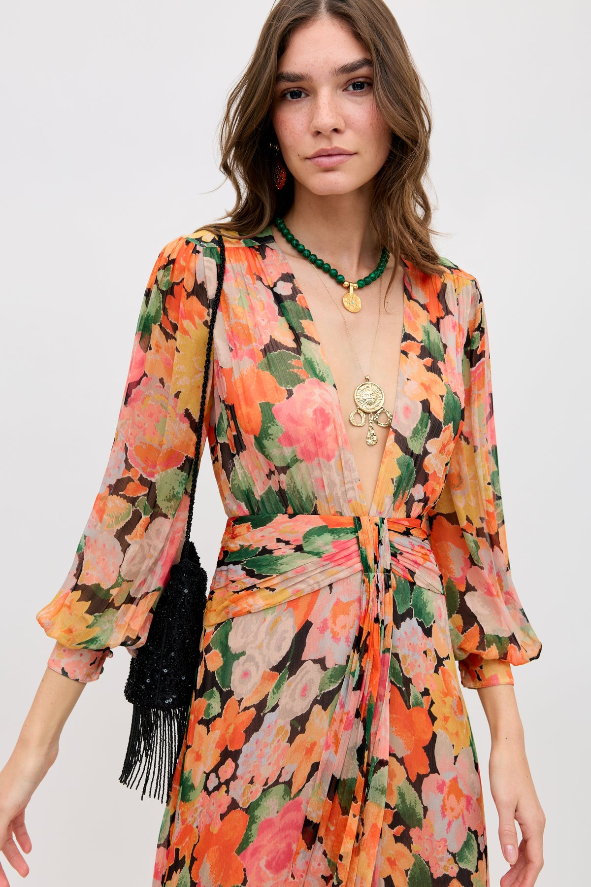 A woman wears a long-sleeved, V-neck floral dress in pink, orange, green, and cream tones. She accessorizes with layered gold necklaces, green beaded jewelry, and the Orla—Vintage Scatter Embellishment Black beaded bag with fringe.