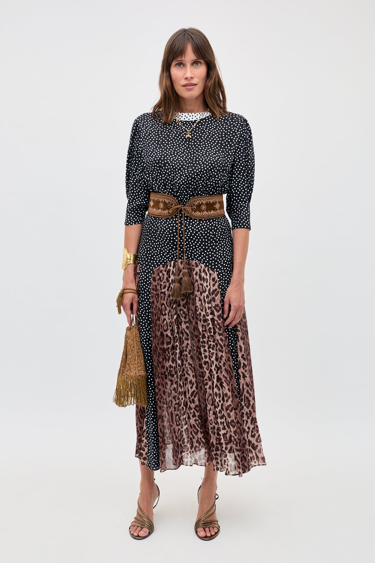 A woman faces forward in the "meg - leopard polka dot" outfit: a black and white polka dot top, leopard print midi skirt, brown velvet belt, fringed gold bag, strappy sandals. She has straight brown hair and a neutral expression.