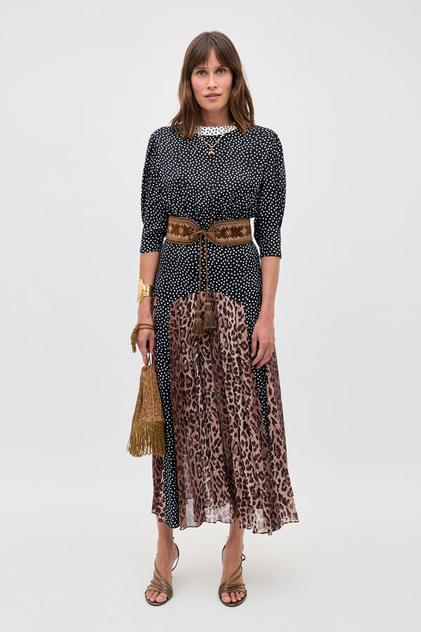 A woman faces forward in the "meg - leopard polka dot" outfit: a black and white polka dot top, leopard print midi skirt, brown velvet belt, fringed gold bag, strappy sandals. She has straight brown hair and a neutral expression.