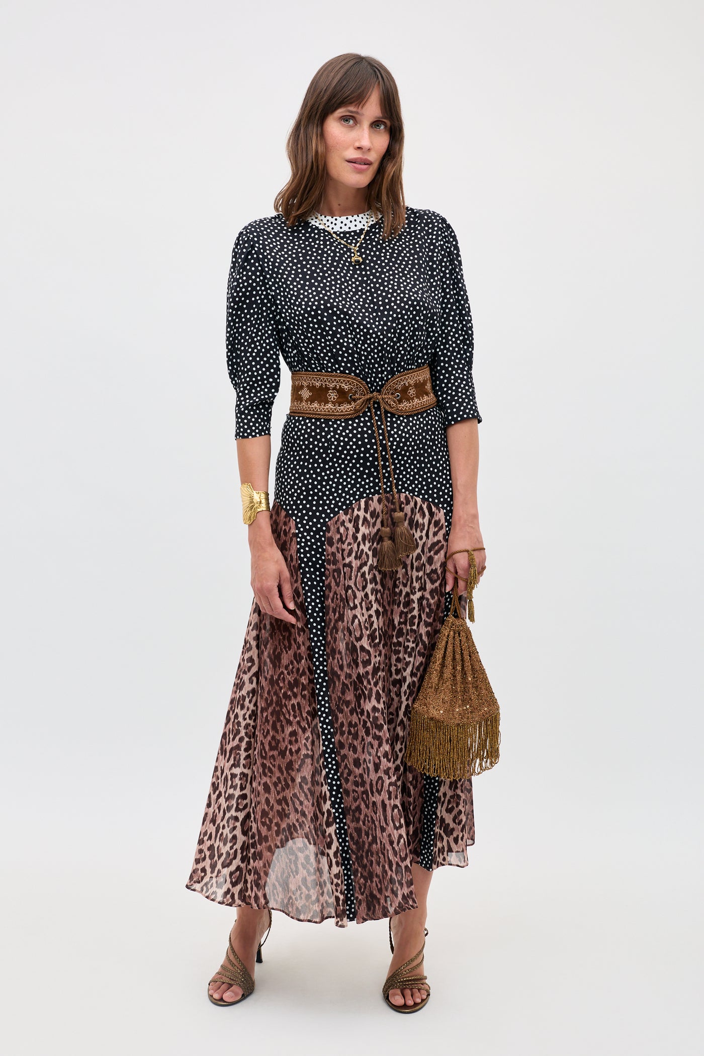 A woman stands against a plain light background wearing the "meg - leopard polka dot" top and skirt, paired with heeled sandals, a wide brown belt, and holding a brown fringed bag.
