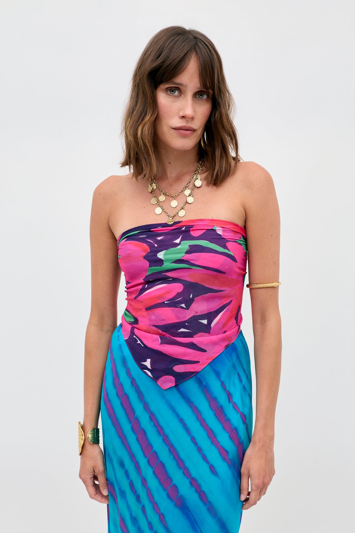 A woman with shoulder-length brown hair wears the Menno - Danish Floral strapless dress, featuring a pink, purple, and blue abstract pattern. She accessorizes with layered gold necklaces, a bangle, and a cuff bracelet against a plain background.