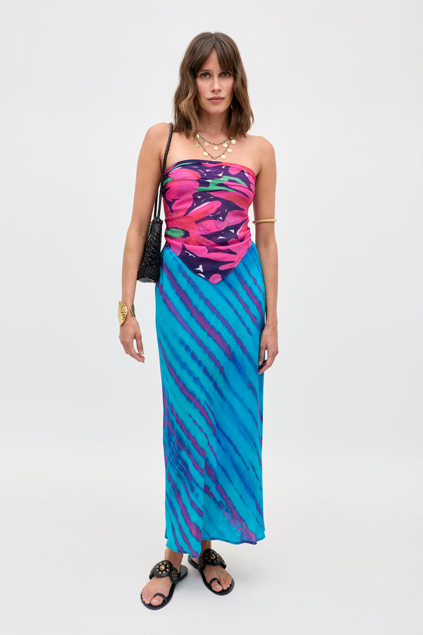A woman poses against a plain background wearing the Menno - Danish Floral dress, featuring a pink floral print top and blue diagonal striped skirt. She accessorizes with a black shoulder bag, layered necklaces, and black sandals.
