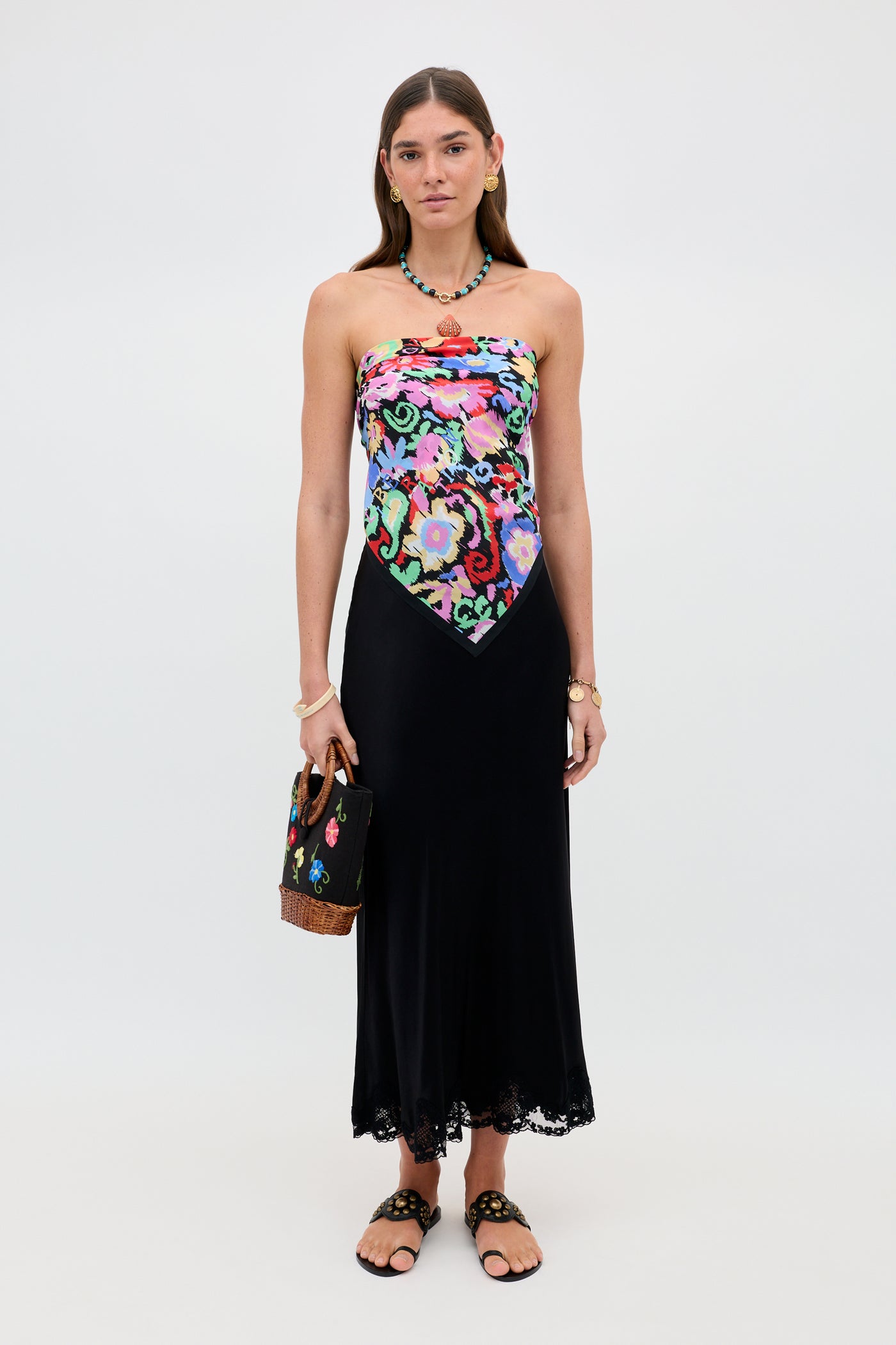 A woman wearing the Menno - Ikat Bloom strapless dress, featuring a vibrant floral bodice and a long black skirt, holds a black handbag with floral embroidery. She accessorizes with sandals, jewelry, and a necklace, standing against a plain white background.