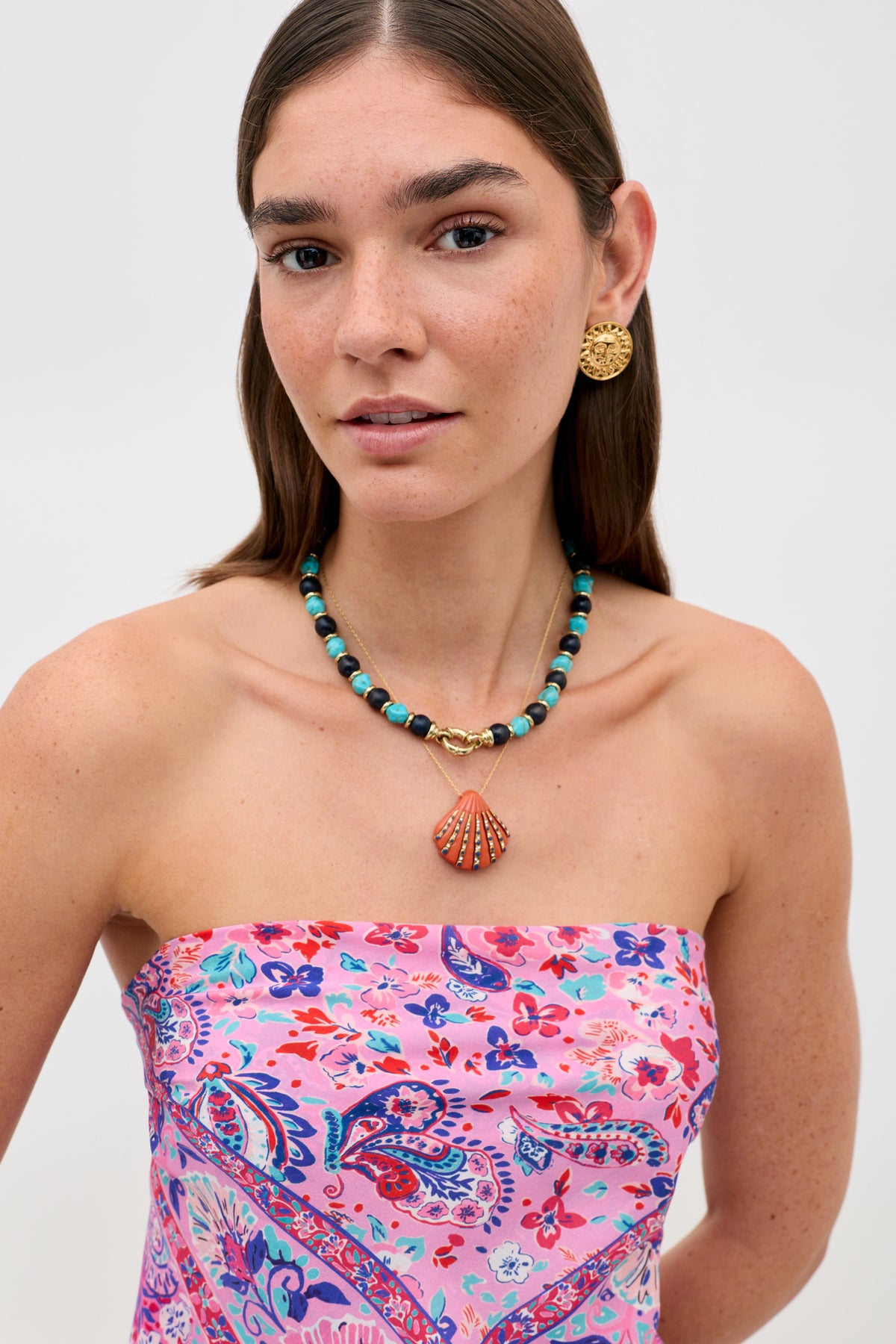 A woman with long brown hair wears Palmara - Blue, a turquoise beaded necklace with a seashell pendant, along with gold statement earrings and a strapless pink floral top. She poses against a plain light background with natural makeup.