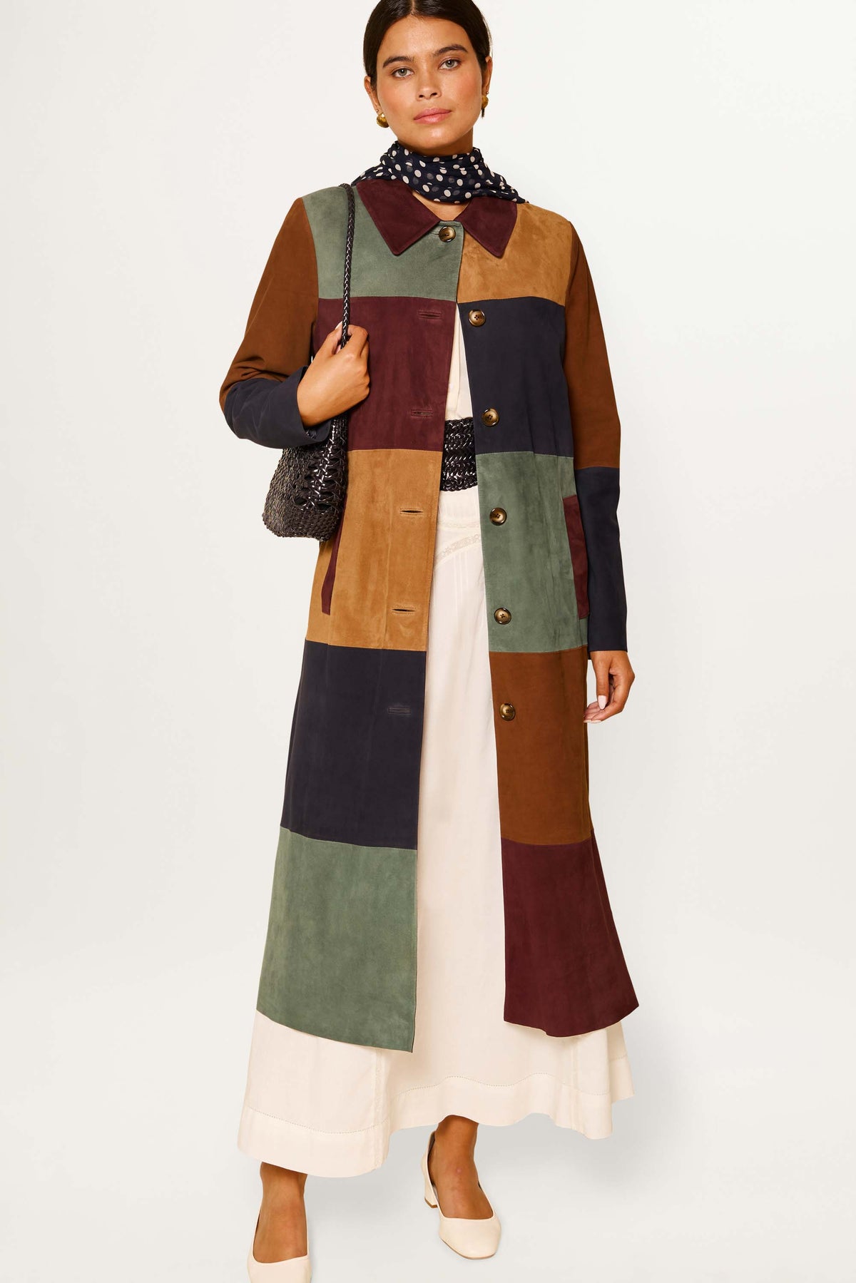 A woman wears the Milly - Patchwork Tan, a long vintage suede coat in earthy tones over a cream skirt. She carries a dark textured bag, has a black and white polka dot scarf, light shoes, and stands against a plain white background.
