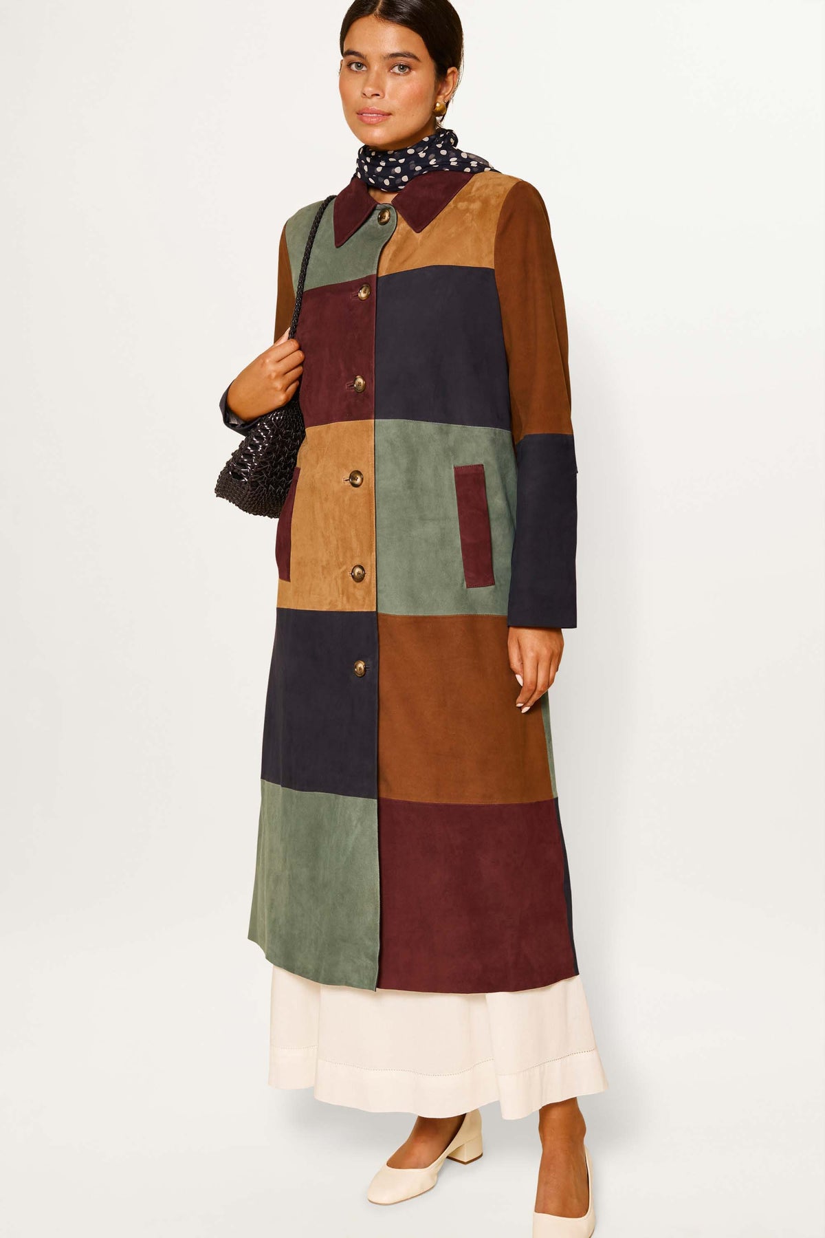 A woman wears the Milly - Patchwork Tan, a long vintage coat in buttery suede with brown, green, navy, and burgundy panels over a white skirt. She holds a textured bag and sports cream shoes against a plain background.