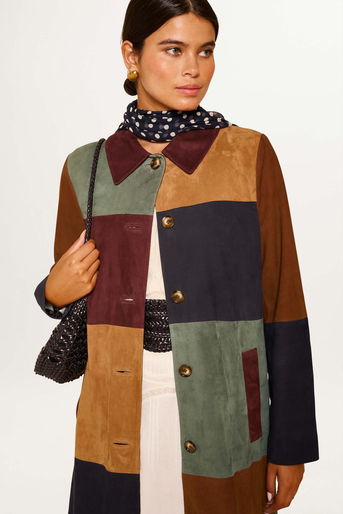 A woman wears the Milly - Patchwork Tan vintage coat in buttery suede with a boxy fit, featuring color-blocks of brown, green, navy, and burgundy. She pairs it with a black and white polka dot scarf and a woven black handbag against a plain background.