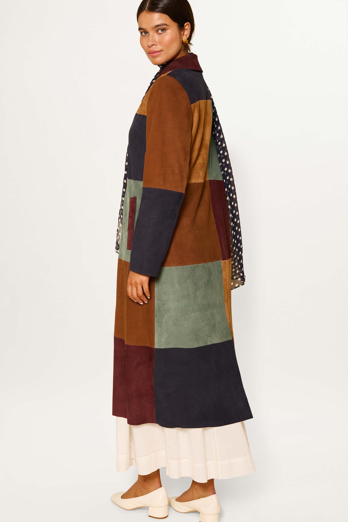 A woman stands sideways wearing the Milly - Patchwork Tan, a vintage-inspired coat in buttery suede earth tones over a cream skirt, with dark hair pulled back and cream shoes against a plain white background.