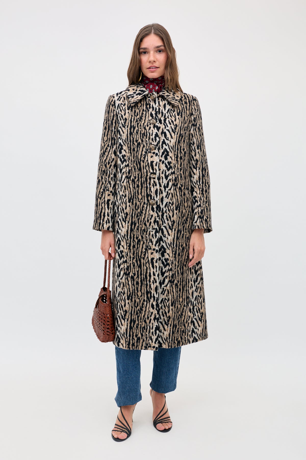 A woman stands against a plain background wearing the Milly - Bohemia Leopard coat over blue jeans and black strappy sandals. She carries a brown woven handbag, her long hair enhancing the effortlessly chic vibe.