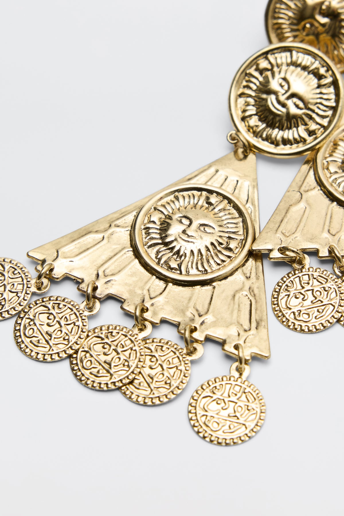 Miray - Gold triangular pendant earrings feature embossed lion faces and circular coin-like charms with intricate patterns, set against a plain light background.