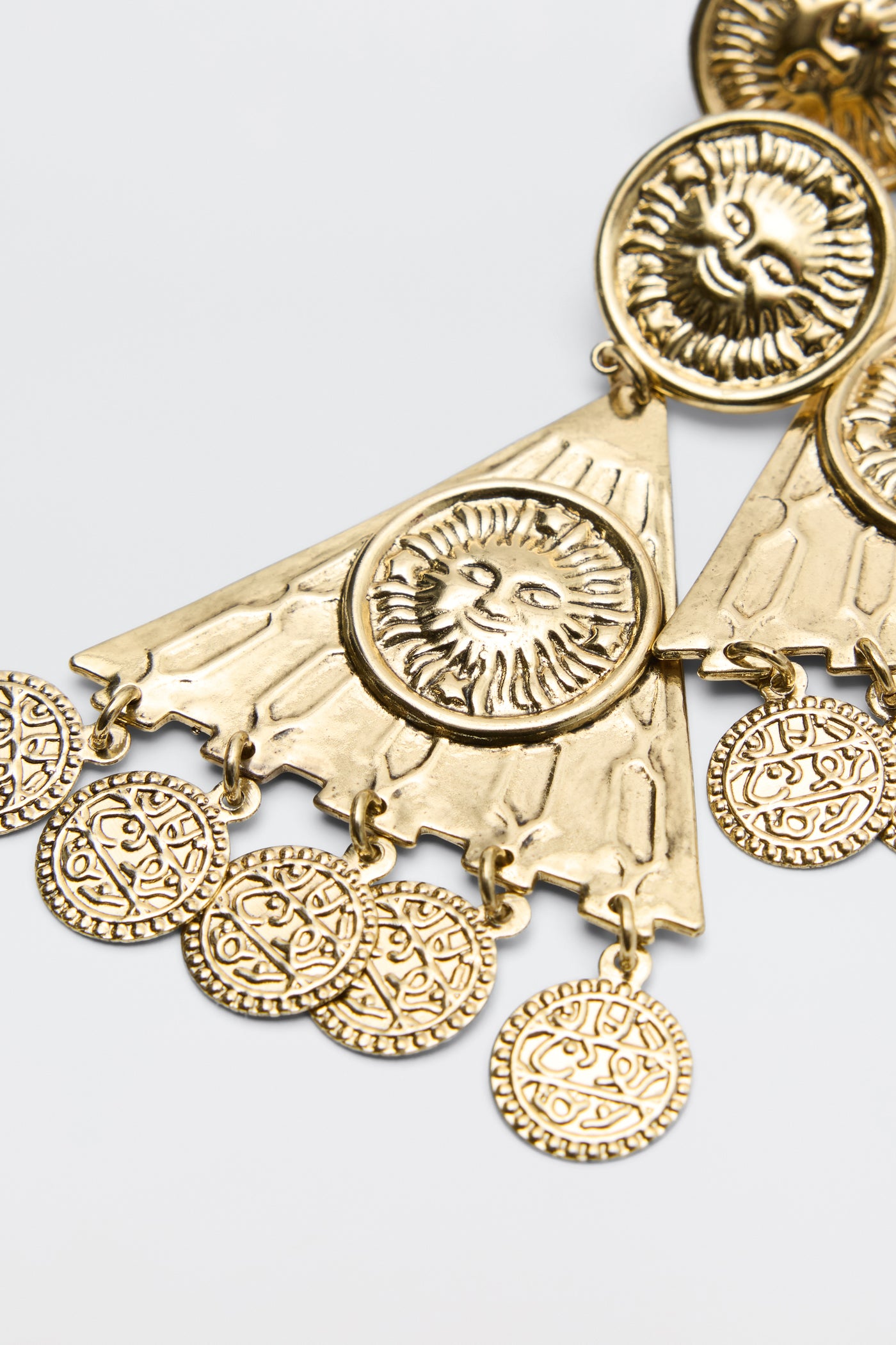 Miray - Gold triangular pendant earrings feature embossed lion faces and circular coin-like charms with intricate patterns, set against a plain light background.