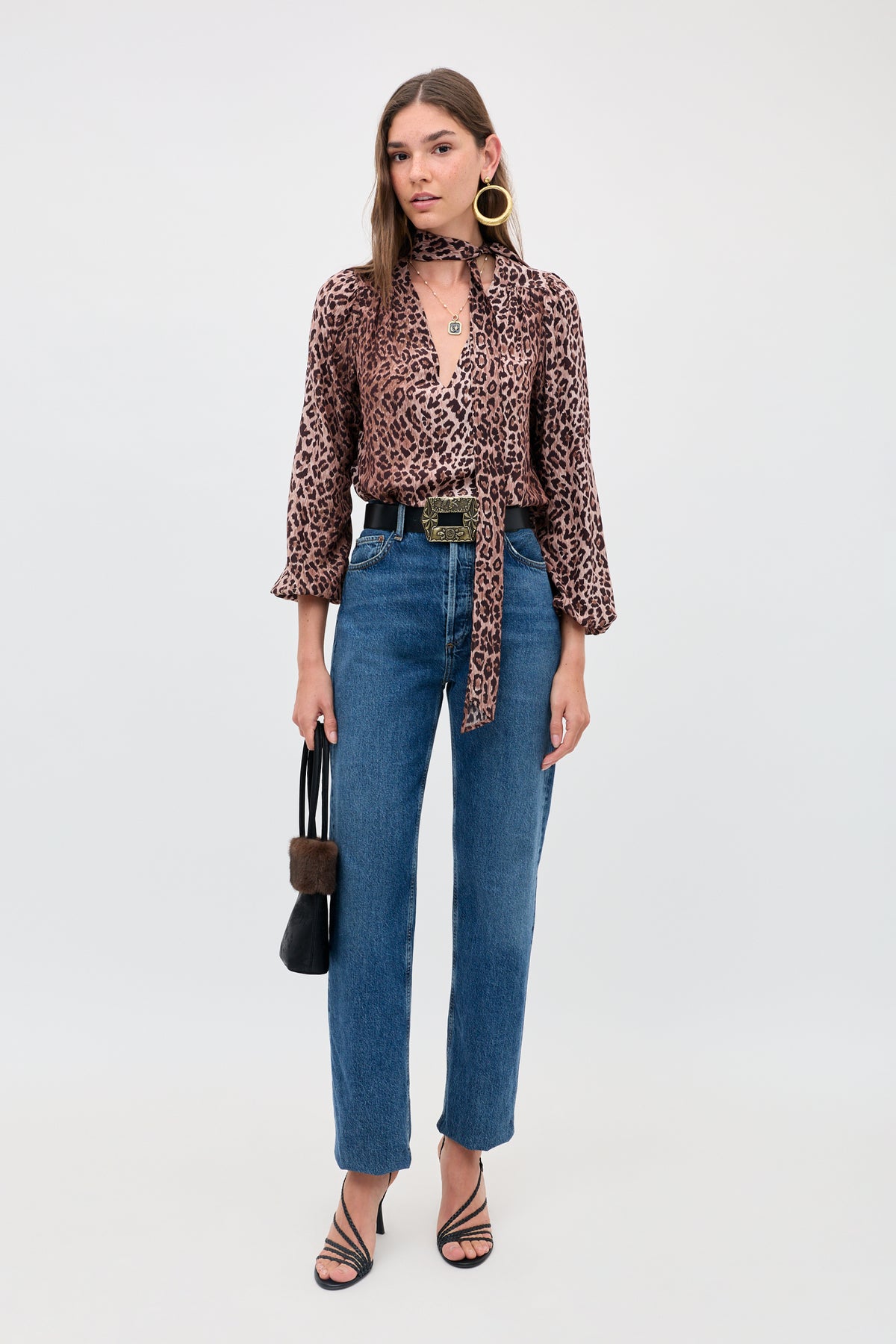 A woman faces forward wearing the Moss - Leopard Brown silk blouse with blouson sleeves and tie, paired with blue high-waisted jeans, black strappy heels, large gold hoop earrings, and holding a black handbag.