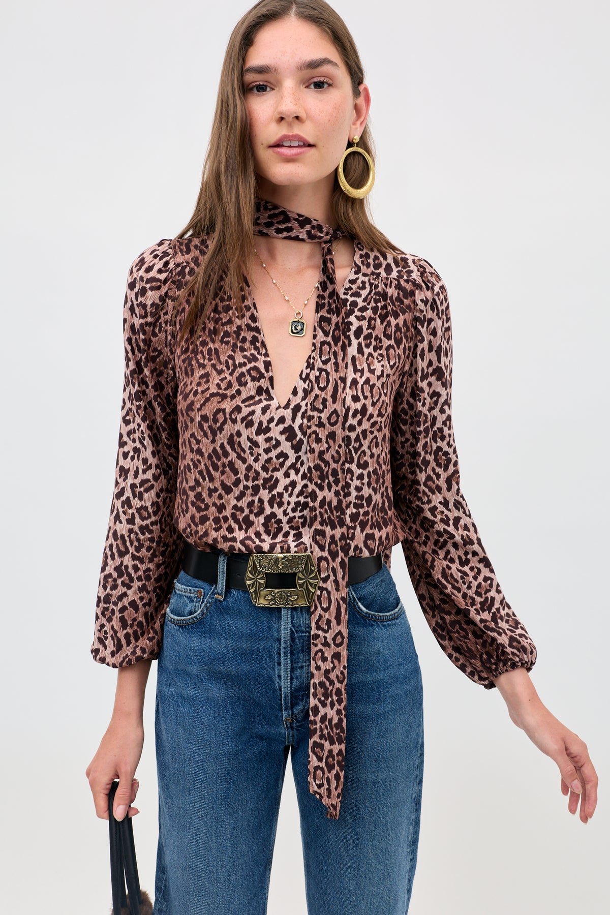 A woman wears the "Moss - Leopard Brown" silk blouse with blouson sleeves and matching scarf, blue jeans with a decorative belt, large hoop earrings, layered necklaces, and holds a black bag against a plain light background.