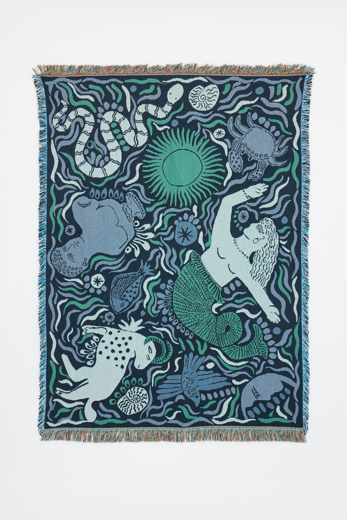 The Mythos Throw - Navy is a woven tapestry featuring motifs inspired by Greek mythology, including a mermaid, a spotted dog, various fish and sea creatures, and a green sun—all set amidst flowing wavy patterns in shades of blue, green, and white. This unique throw is finished with fringed edges.
