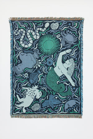 The Mythos Throw - Navy is a woven tapestry featuring motifs inspired by Greek mythology, including a mermaid, a spotted dog, various fish and sea creatures, and a green sun—all set amidst flowing wavy patterns in shades of blue, green, and white. This unique throw is finished with fringed edges.
