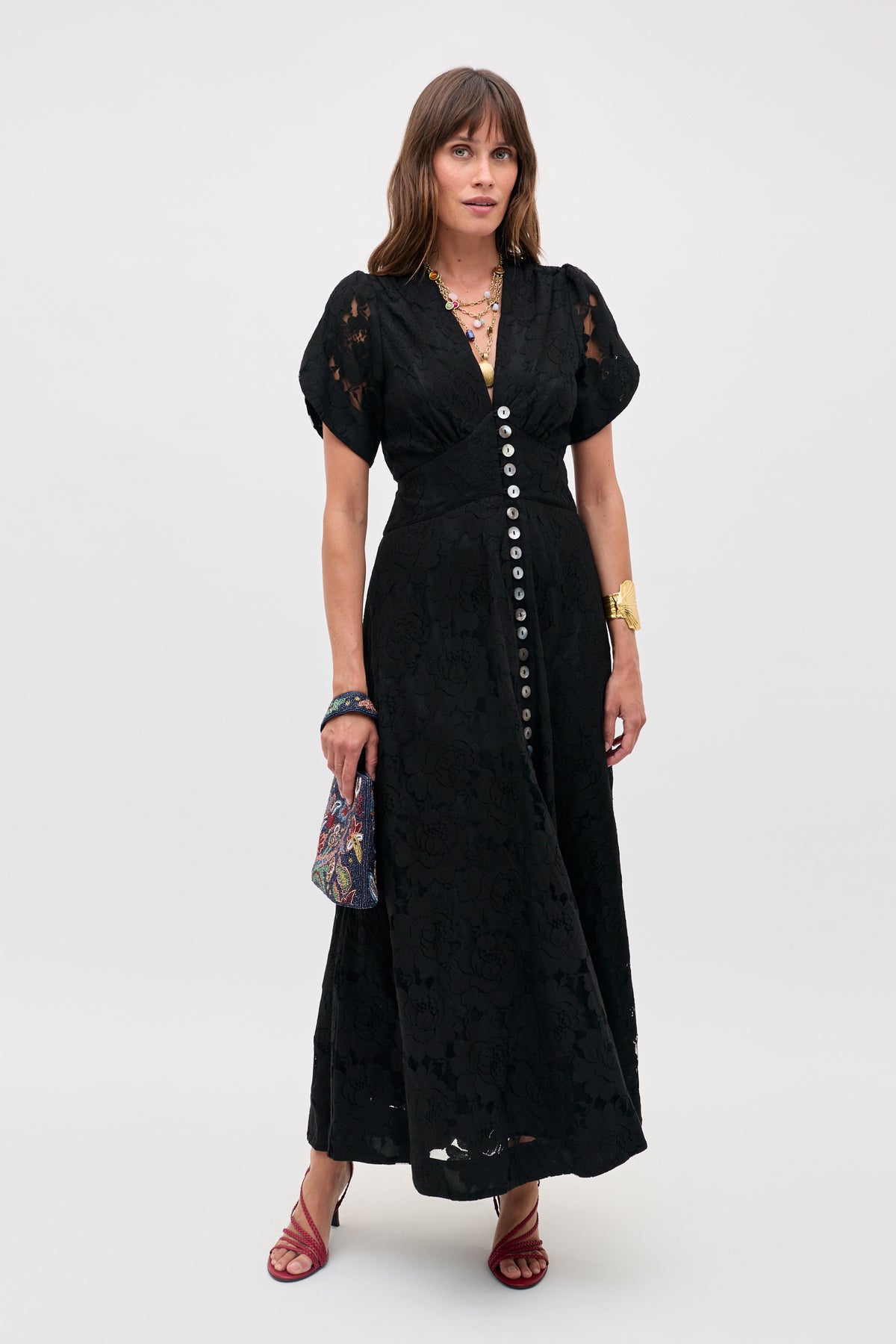 A woman in the Novara - Black short-sleeved, button-down lace dress with red strappy sandals, gold jewelry, and a patterned handbag stands against a plain light background.