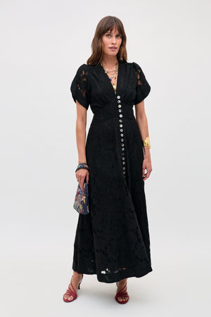 A woman in the Novara - Black short-sleeved, button-down lace dress with red strappy sandals, gold jewelry, and a patterned handbag stands against a plain light background.