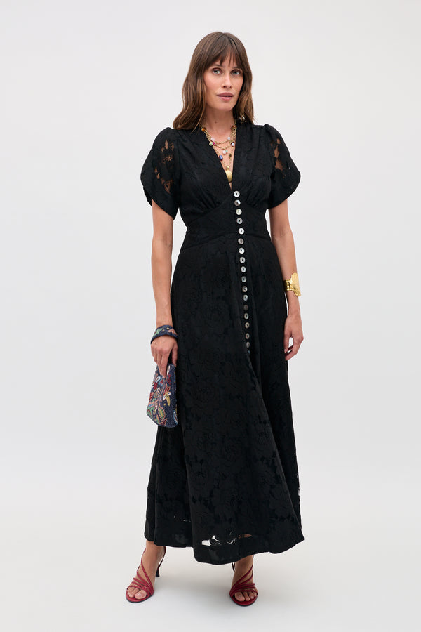 A woman in the Novara - Black short-sleeved, button-down lace dress with red strappy sandals, gold jewelry, and a patterned handbag stands against a plain light background.