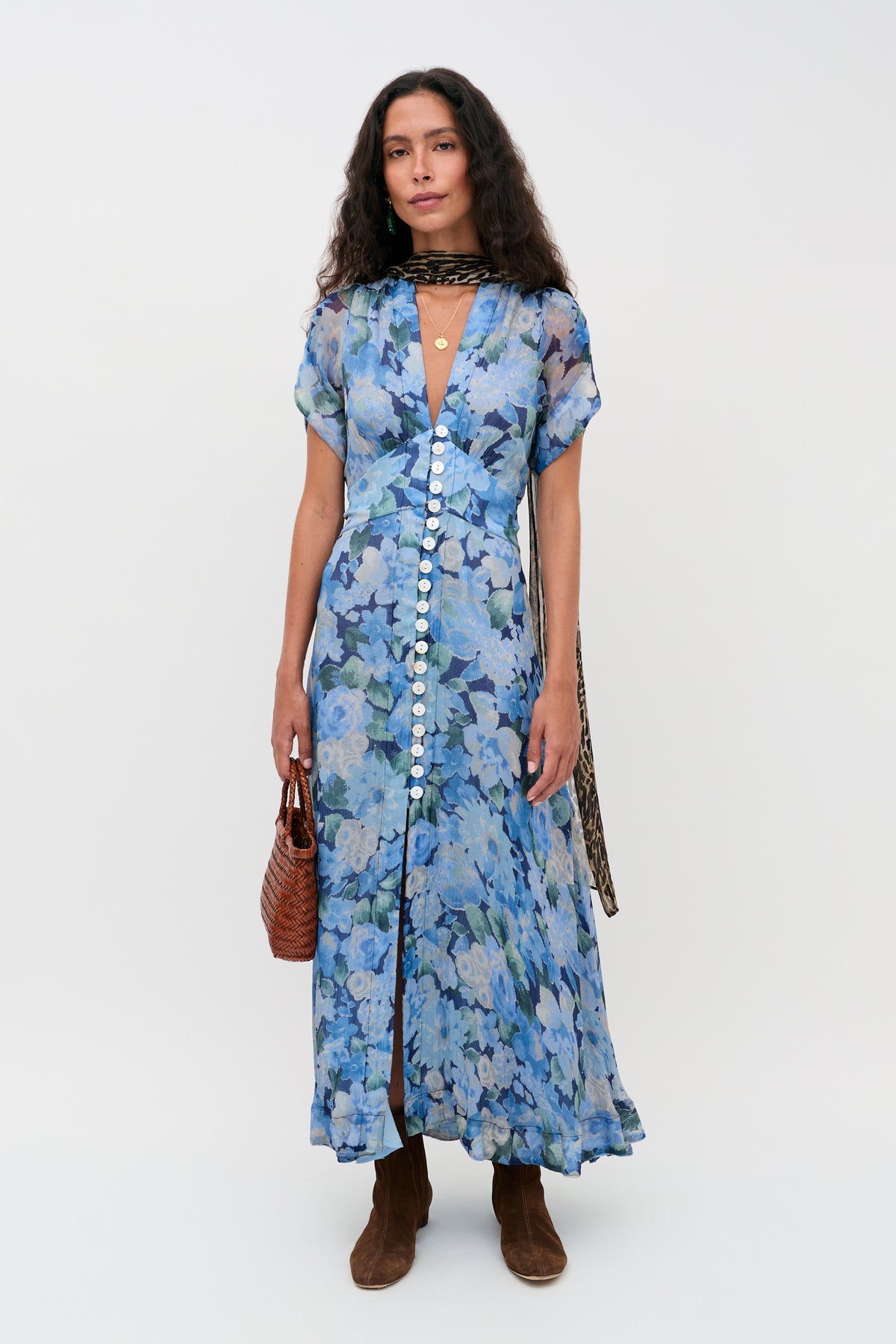 A woman wears the Novara Dress in Chelsea Floral Blue, styled with brown boots, a brown woven handbag, and a patterned scarf around her neck, standing against a plain white background.