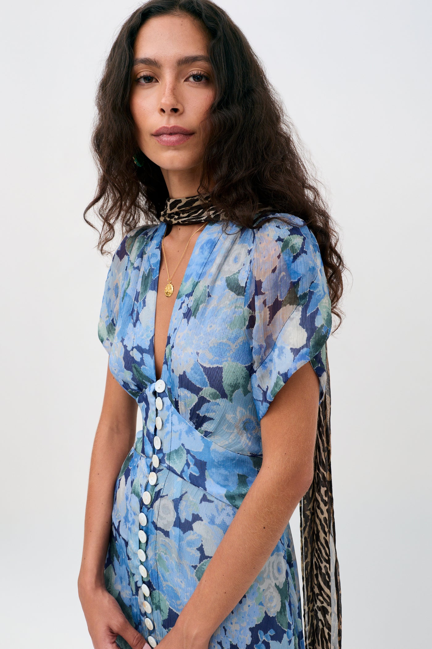 A woman with long wavy brown hair is wearing the Novara Dress in Chelsea Floral Blue, featuring white buttons, paired with a leopard print scarf, standing against a plain light background.