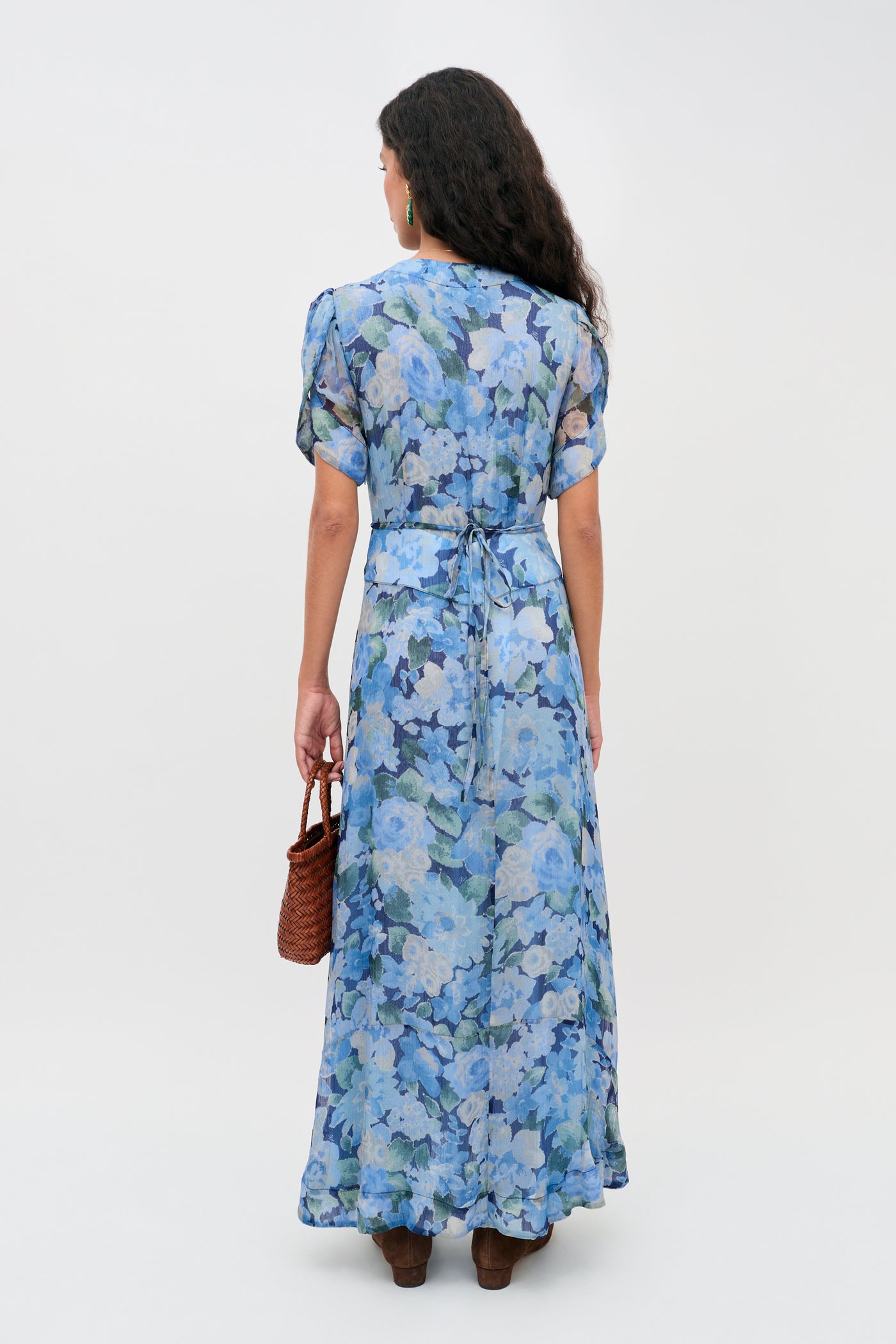 A woman with long dark hair stands facing away, wearing the Novara Dress in Chelsea Floral Blue, featuring short sleeves and a waist tie. She accessorizes with a brown woven handbag and brown shoes, posing against a plain white background.