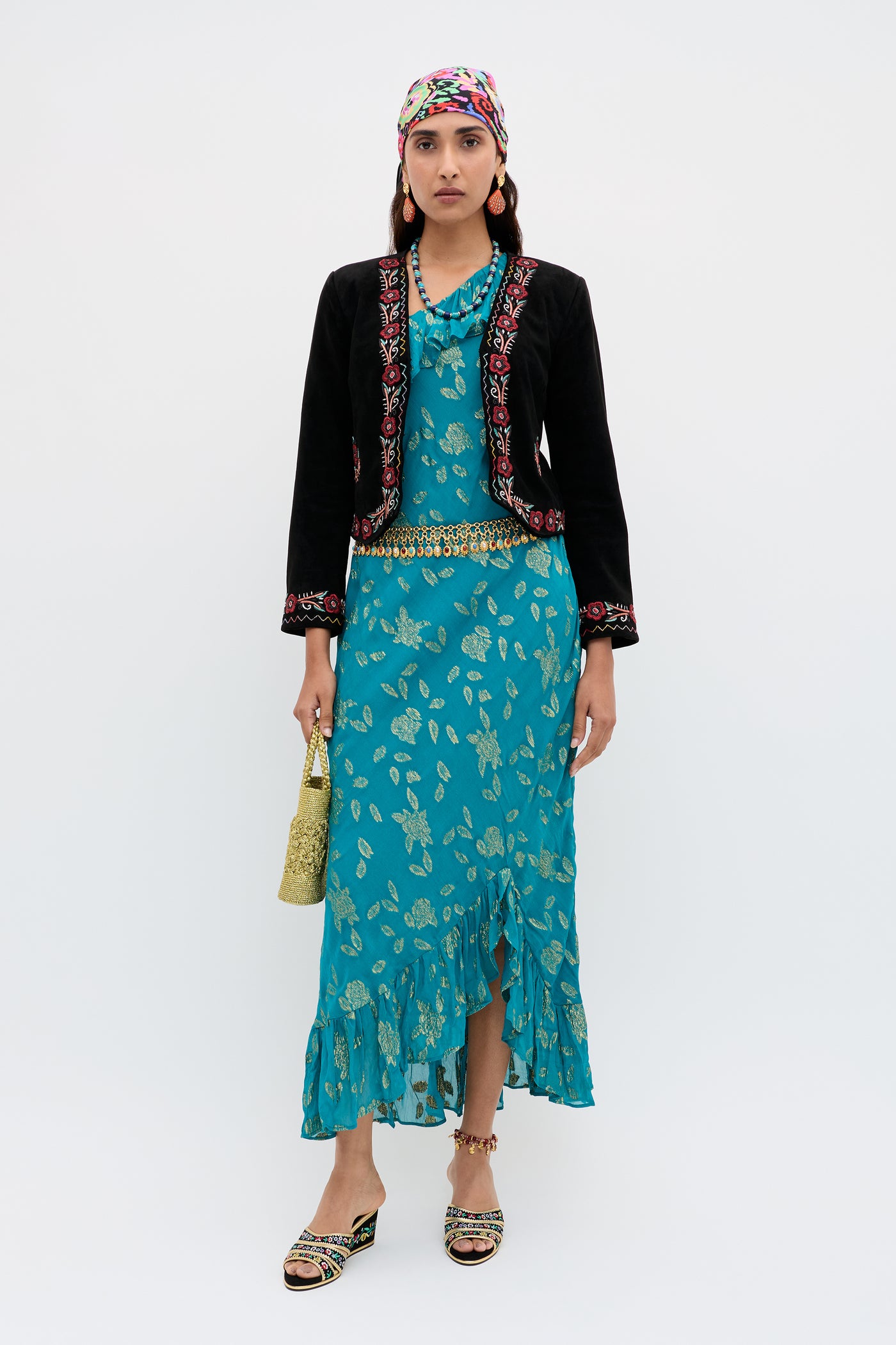 A woman stands against a plain white background wearing the Oliwia - Roselle Embellishment Black jacket, styled with a patterned headscarf, teal printed dress, gold belt, sandals, and holding a gold handbag.
