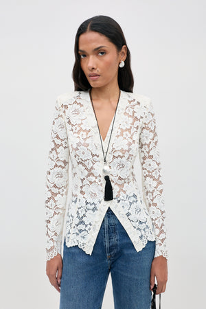 A woman wearing the Padma - Cream white lace long-sleeve top and blue jeans stands against a plain background, accessorized with a black tassel necklace and round earrings.