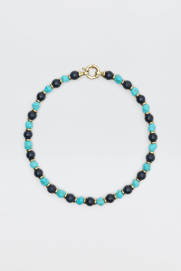 The "Palmara - Blue" necklace features alternating blue-green and dark blue round beads with gold spacers and a gold spring ring clasp, elegantly displayed against a plain light background.