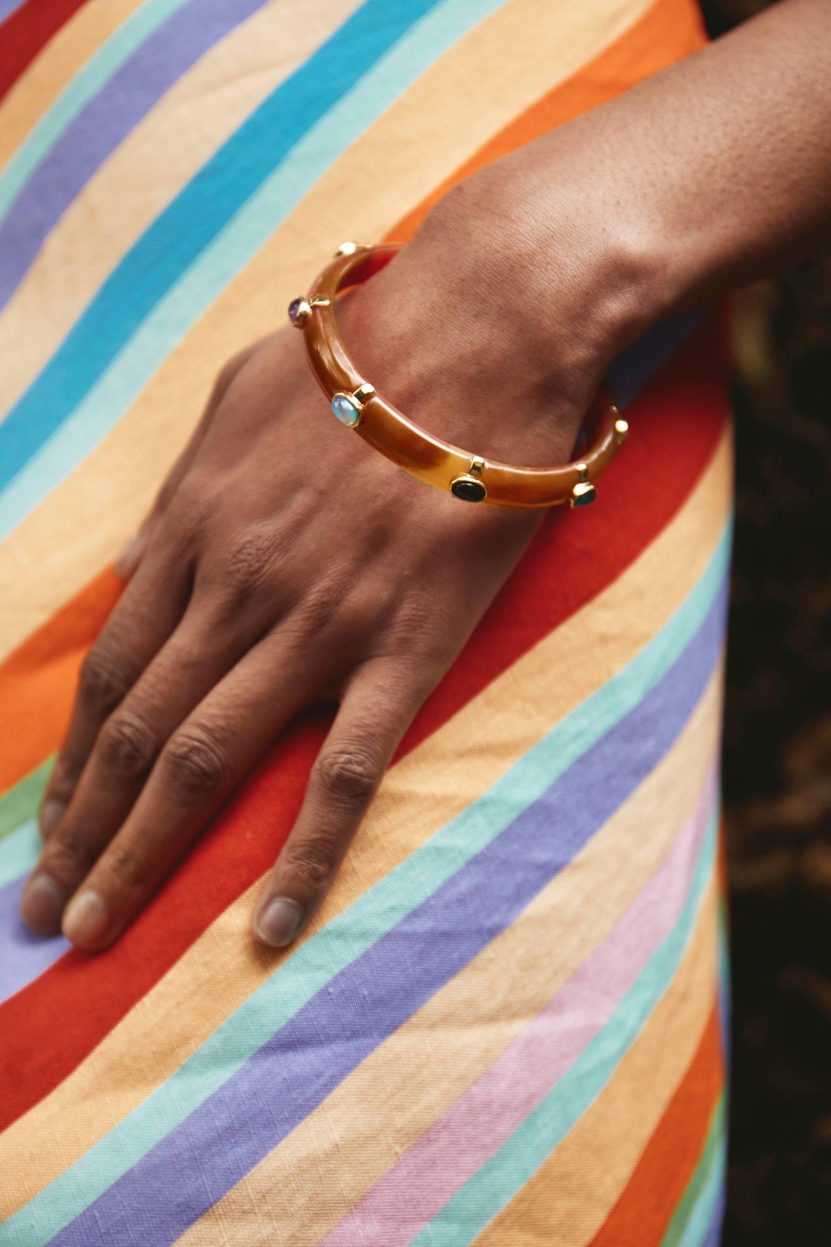 A hand adorned with the Sabella - Tortoise bangle featuring multicoloured cabochons rests on a dress with bold diagonal stripes in orange, yellow, blue, purple, and green.