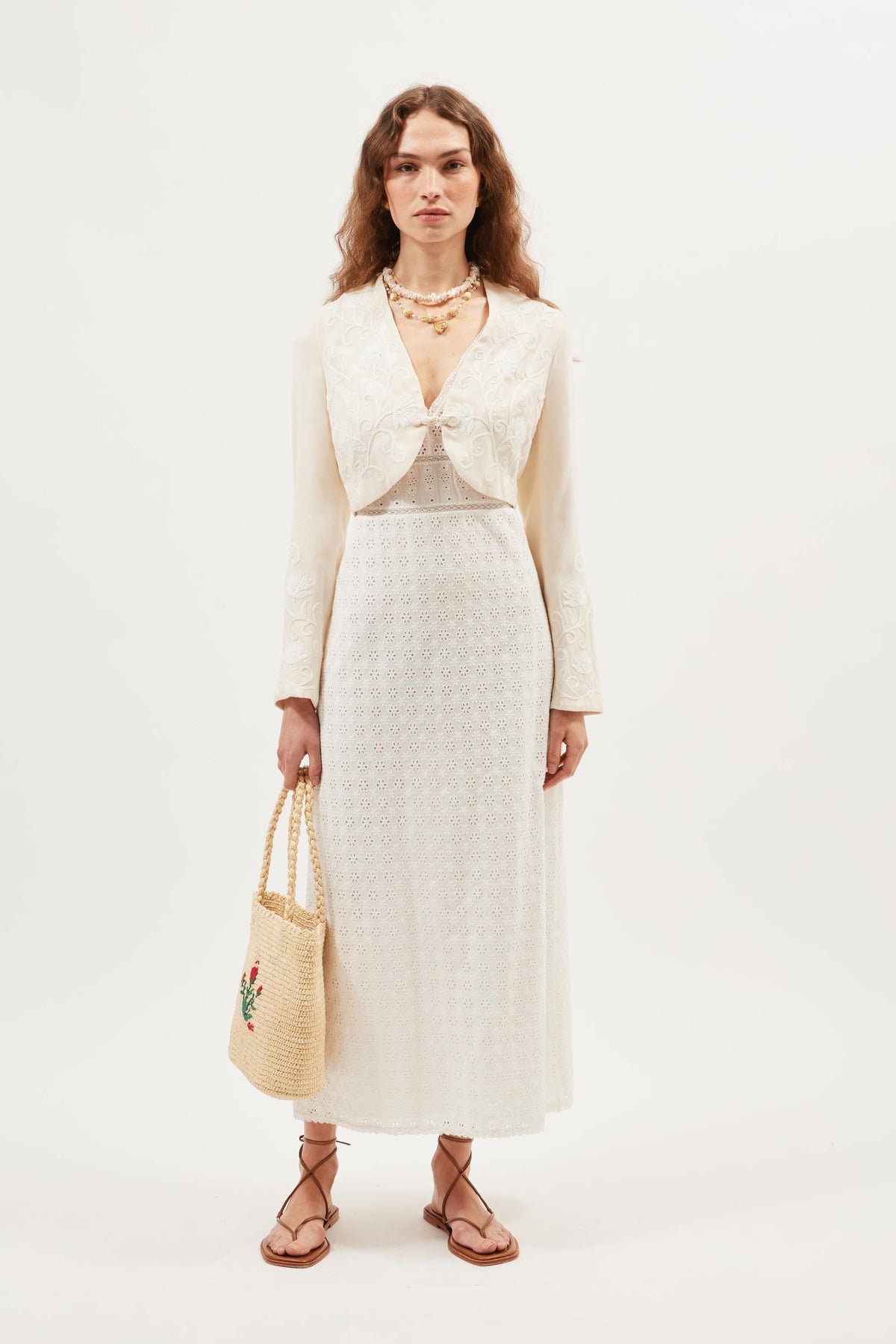 A woman stands against a white background wearing the Learke - Cream, a long, white embroidered dress. She pairs it with layered necklaces, flat sandals, and holds a floral-embroidered woven straw bag.