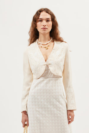 A woman wears the Learke - Cream vintage souvenir jacket with floral embroidery over a white eyelet dress, accessorized with layered pearl necklaces, standing against a plain white background.