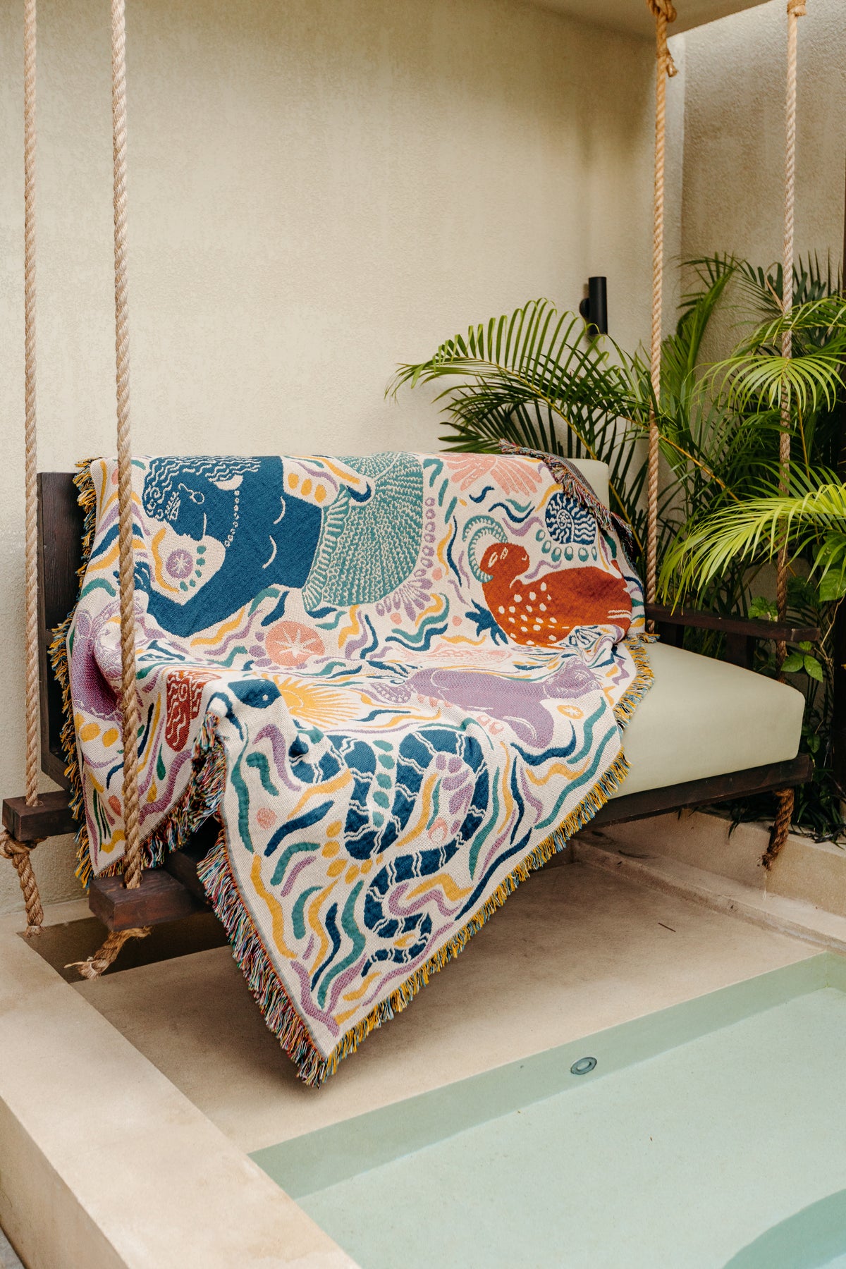 The Athena Throw - Ivory, a recycled cotton piece from the Mythos Collection inspired by Greek mythology, adds color and pattern to a cushioned swing seat on a patio with potted plants by the pool.
