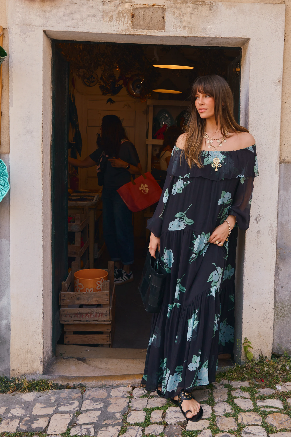 A woman wearing the Lela - Peonies Scatter Charcoal off-the-shoulder dress stands in a doorway holding a black bag. Inside the shop, another person is partially visible with a red tote bag, and cobblestone pavement stretches across the foreground.