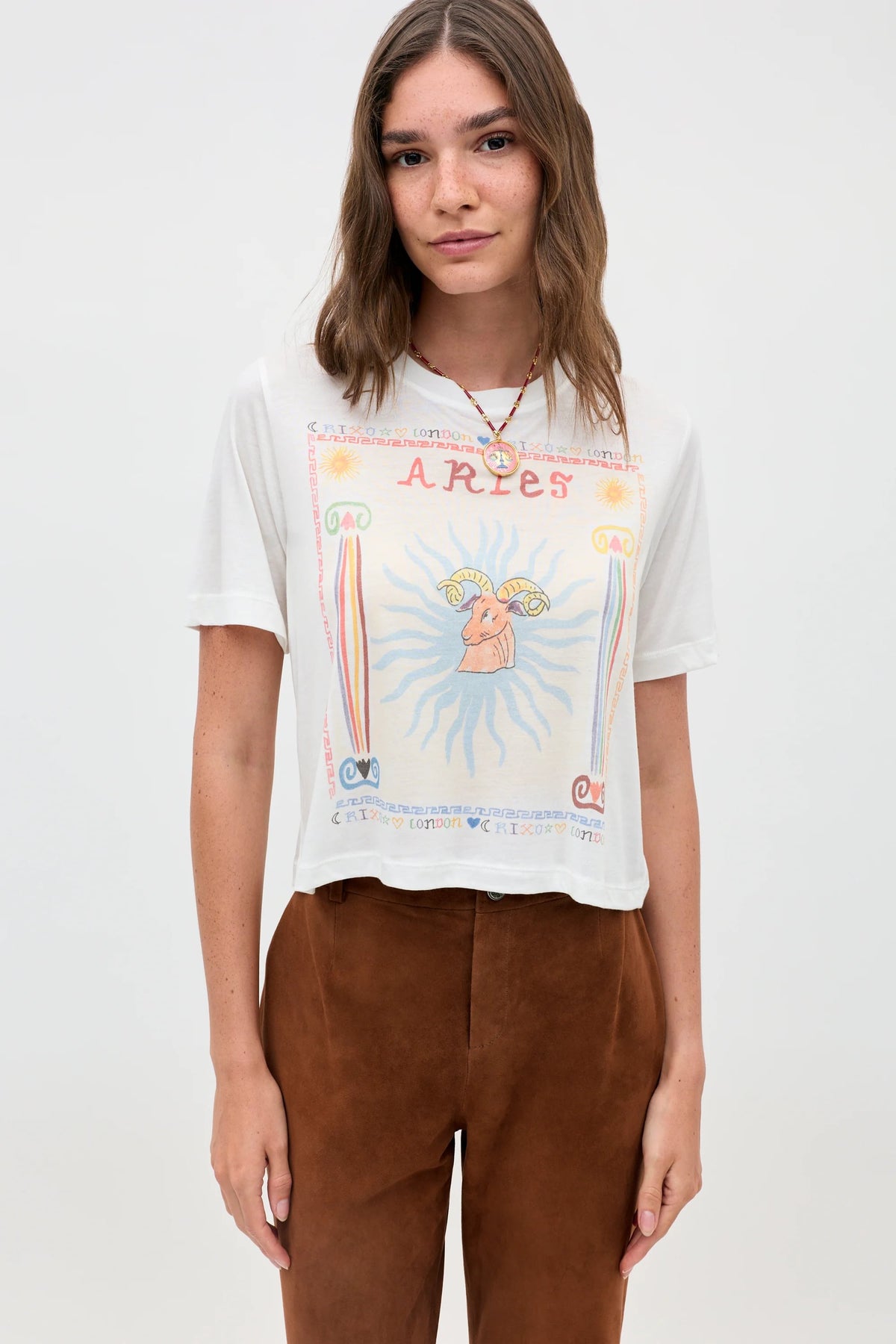 A woman with long brown hair wears the Ria - Aries white sheer jersey T-shirt with an Aries illustration and brown pants, standing against a plain light background.
