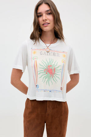 A woman with long brown hair stands against a plain light background, wearing the Ria - Cancer t-shirt. The white top features a vibrant Cancer zodiac sign graphic with a bold red crab illustration and a cropped hem, paired with brown pants.