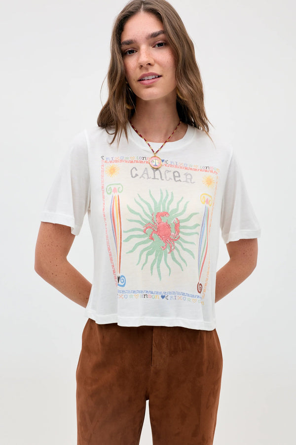A woman with long brown hair stands against a plain light background, wearing the Ria - Cancer t-shirt. The white top features a vibrant Cancer zodiac sign graphic with a bold red crab illustration and a cropped hem, paired with brown pants.