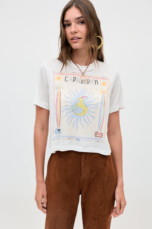 A woman wearing the Ria - Capricorn white T-shirt, featuring a vibrant Capricorn illustration, paired with brown pants and standing against a plain light background.