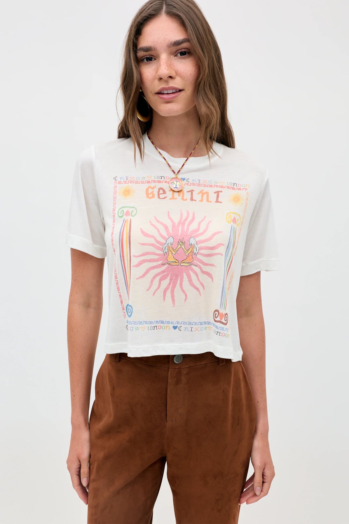 A woman stands facing the camera against a plain light background, wearing the "Ria - Gemini" white sheer jersey t-shirt featuring a colorful Gemini illustration and paired with brown pants.