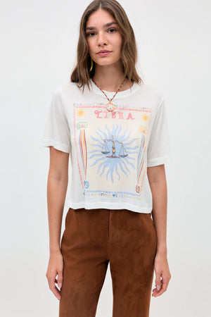 A woman with straight, light brown hair wears the Ria - Libra cropped white T-shirt, featuring scales and sun rays, paired with brown pants. She stands against a plain light background, highlighting the tee’s modern cut.