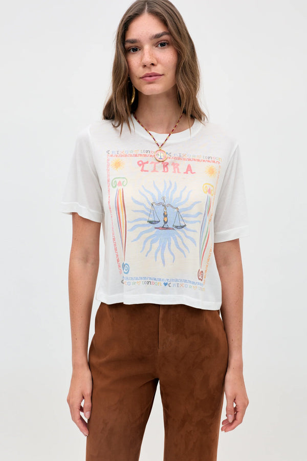 A woman with straight, light brown hair wears the Ria - Libra cropped white T-shirt, featuring scales and sun rays, paired with brown pants. She stands against a plain light background, highlighting the tee’s modern cut.