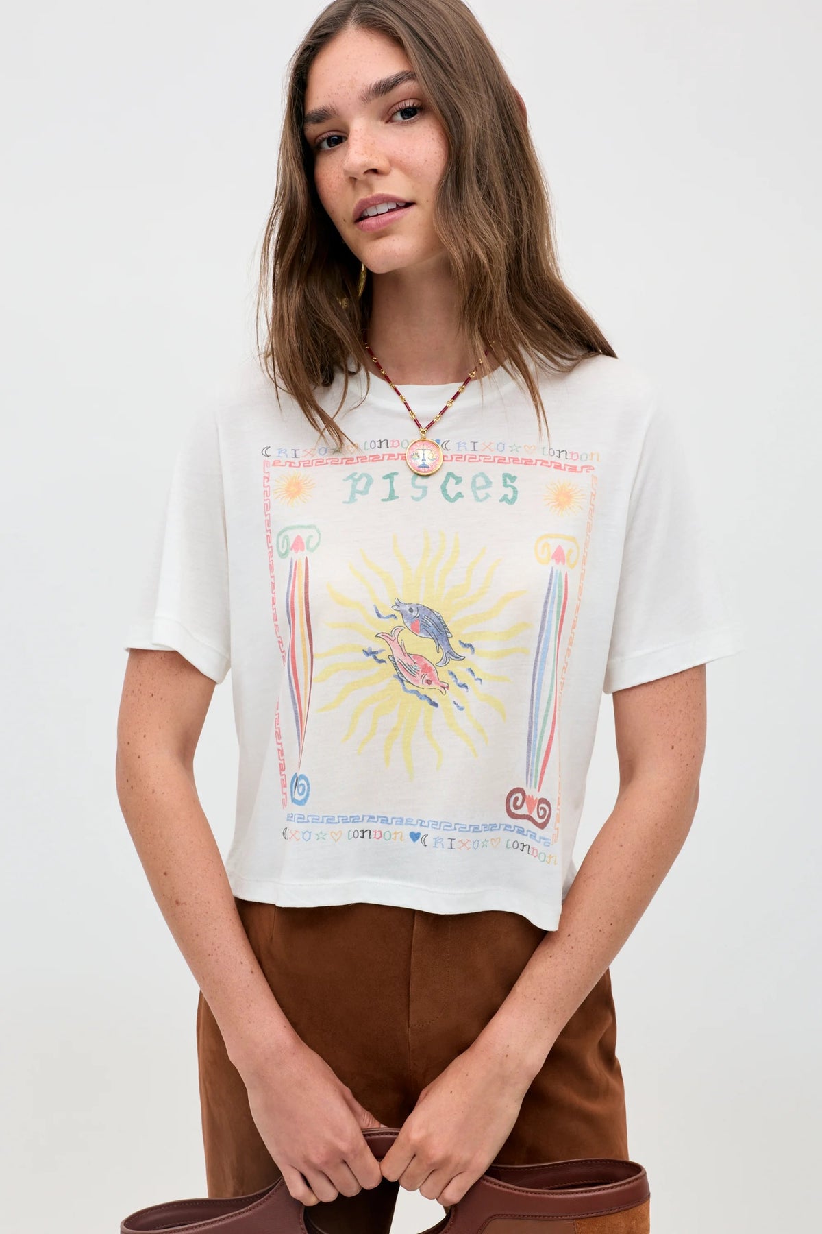 A woman with long brown hair wears the "Ria - Pisces" white T-shirt, featuring a vibrant sun and fish illustration; she pairs it with brown pants and a necklace, standing against a plain light background.